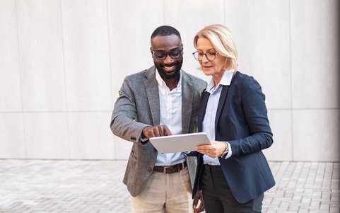 Zwei Geschäftsleute stehen vor einem Gebäude und schauen gemeinsam auf ein Tablet.