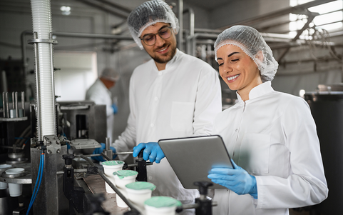 Zwei Mitarbeitende in weißen Kitteln und Haarnetzen schauen auf ein Tablet an einer Lebensmittel-Produktionslinie.